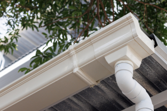 Gutter White plastic rain gutter on a roof edge with downspout, ideal for Colorado Springs roofing or home exterior restoration; tree branches behind.