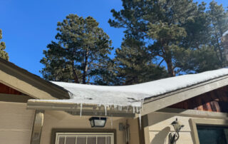 Snow and long icicles hang from a roof, highlighting the need for exterior home restoration in Colorado Springs; evergreens behind.