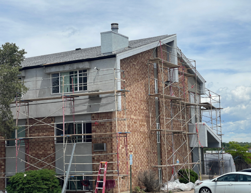 When Is Replacement the Right Call A two-story brick building in Colorado Springs with scaffolding for roofing or exterior restoration; a white car is parked nearby.
