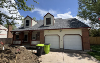 roofing company Colorado Springs A two-story house in Colorado Springs with a front porch, garage doors, dormer windows—roof restoration underway, bins by dirt pile.